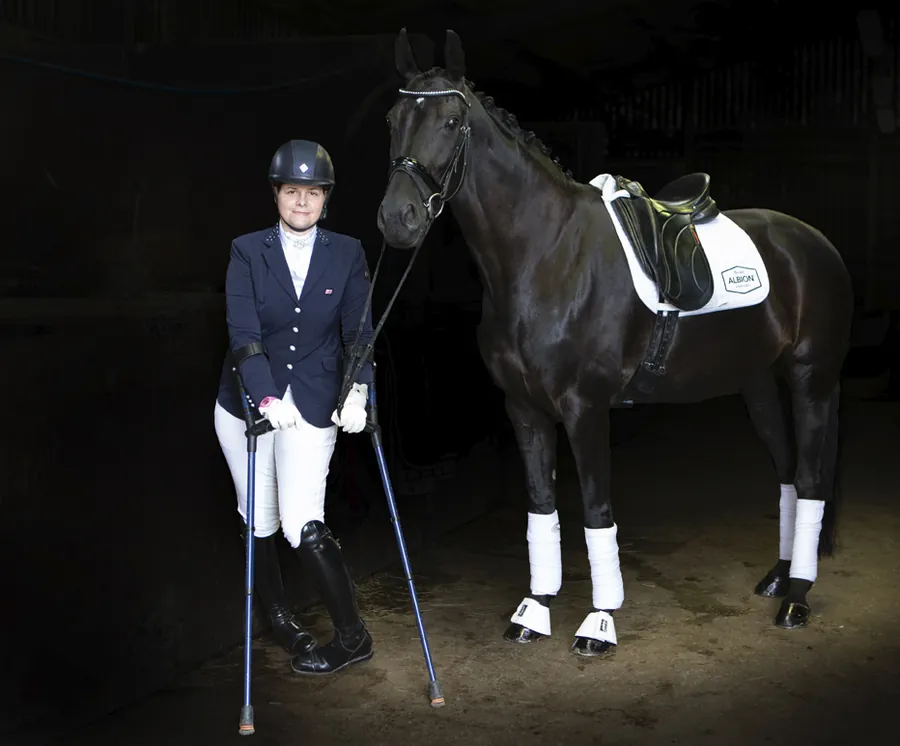 Amanda in show jacket and white breeches standing with crutches beside her black horse