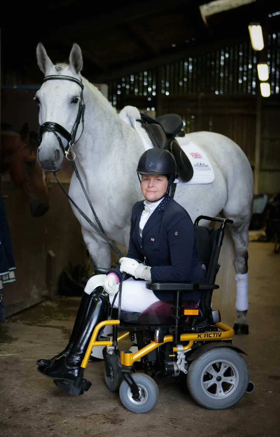Amanda in competition dress in her wheelchair in the stable yard, with her grey mare Ruby Rose II tacked up behind her