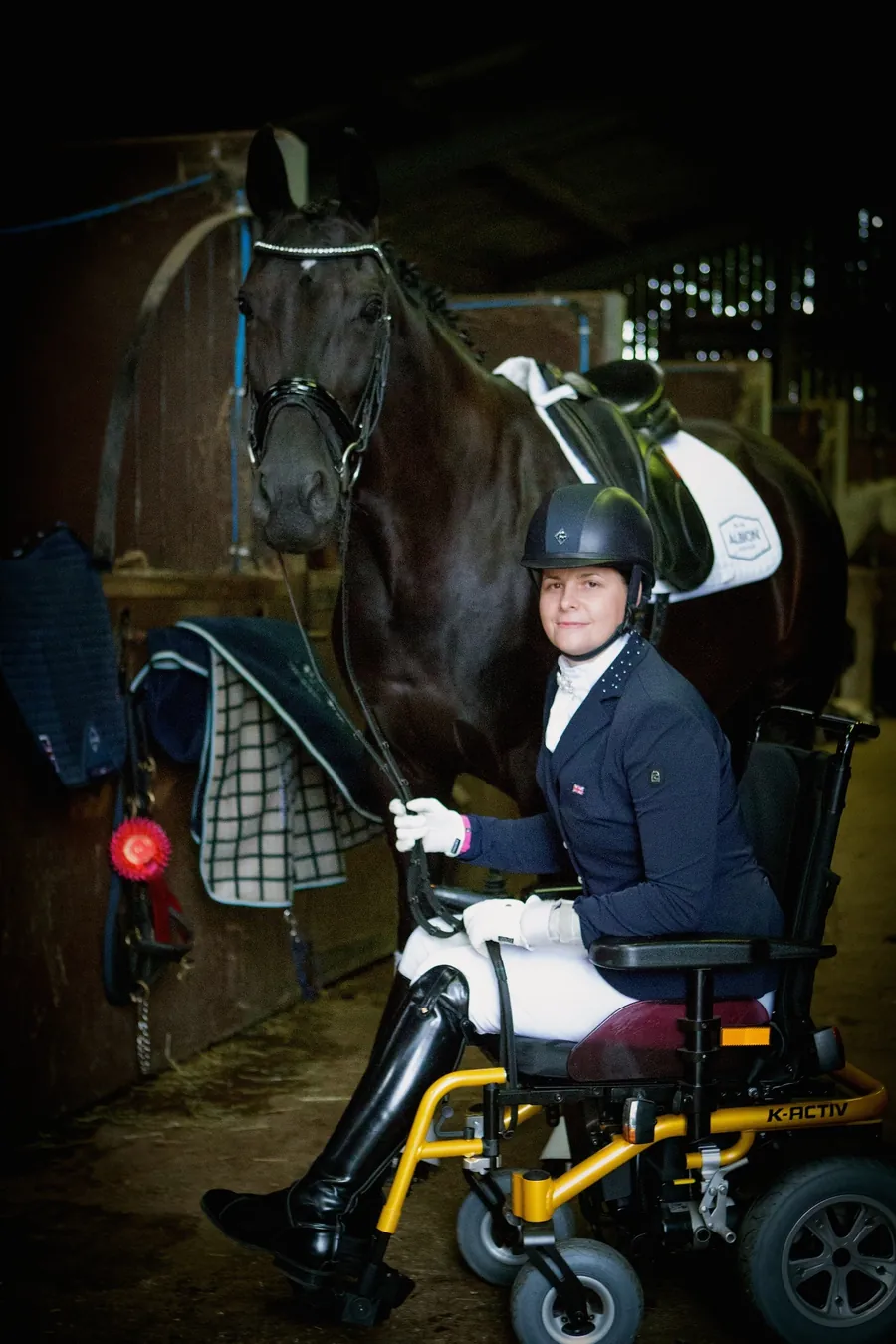 Amanda in competition dress in her wheelchair, holding her black horse in the stable yard
