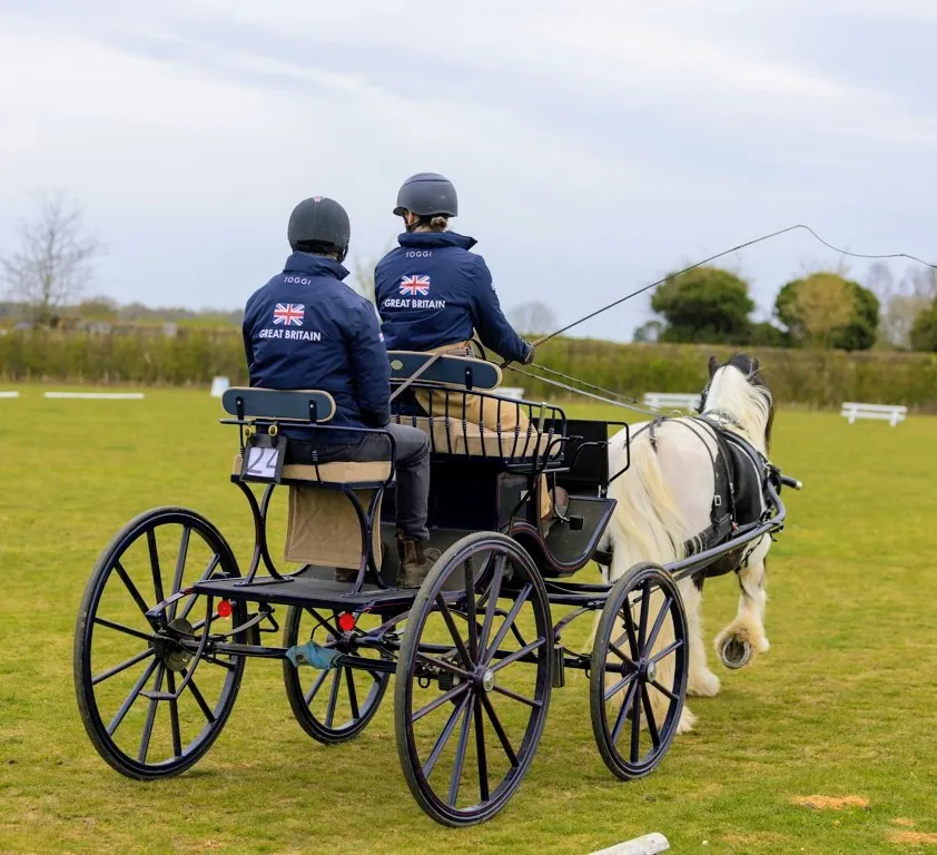 Amanda and her back-stepper in Great Britain jackets on her carriage behind a skewbald cob, seen from behind at a show