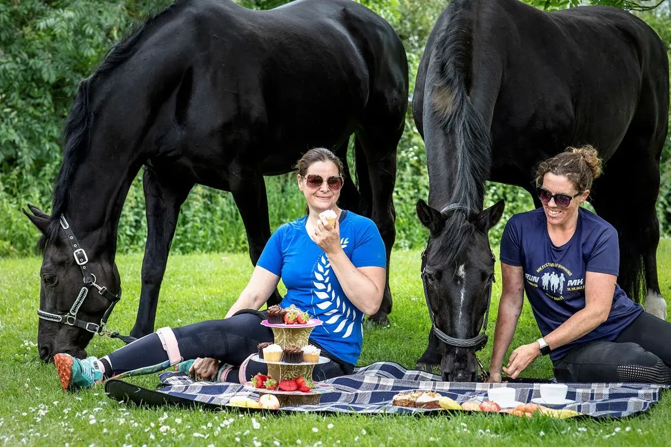 Two women enjoying a cake picnic on the grass beside two grazing black horses for Graze and Gather for Good