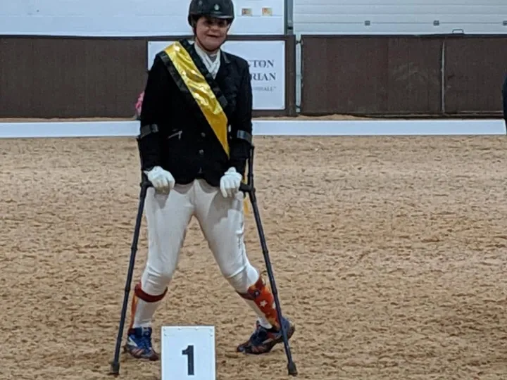 Amanda on crutches in a yellow champion’s sash beside a number 1 podium marker in an indoor arena