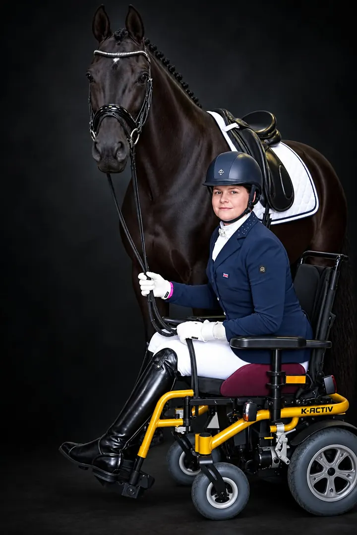 Amanda in show jacket and riding hat in her wheelchair, holding a black dressage horse, in a studio portrait