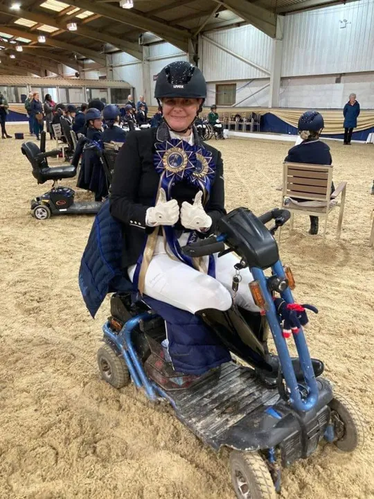 Amanda on her mobility scooter in an indoor arena, wearing two blue championship sashes and giving two thumbs up