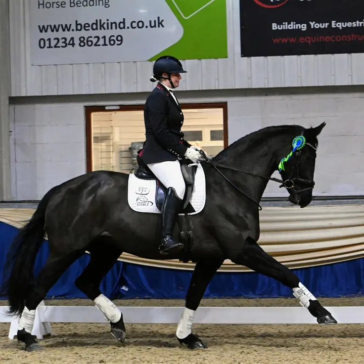 Amanda riding a black horse in an indoor arena with a Country & Stable saddle pad and a rosette on the bridle