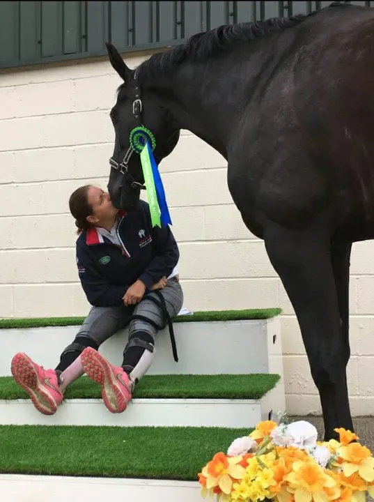 Amanda sitting on steps kissing her black horse, who wears a winner’s rosette