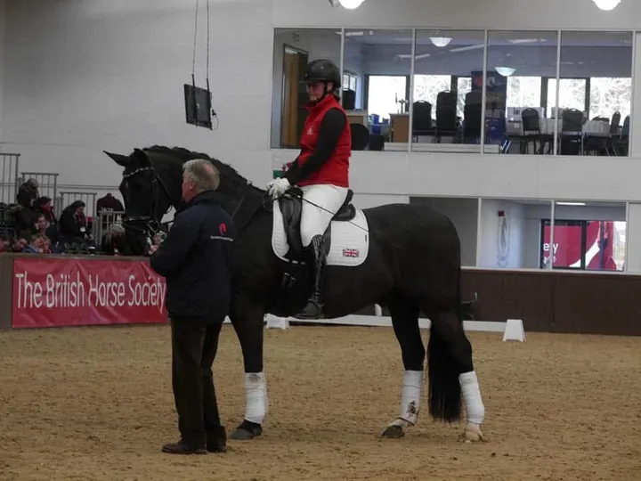 Amanda on a black horse beside a coach in a British Horse Society arena, with spectators in the gallery