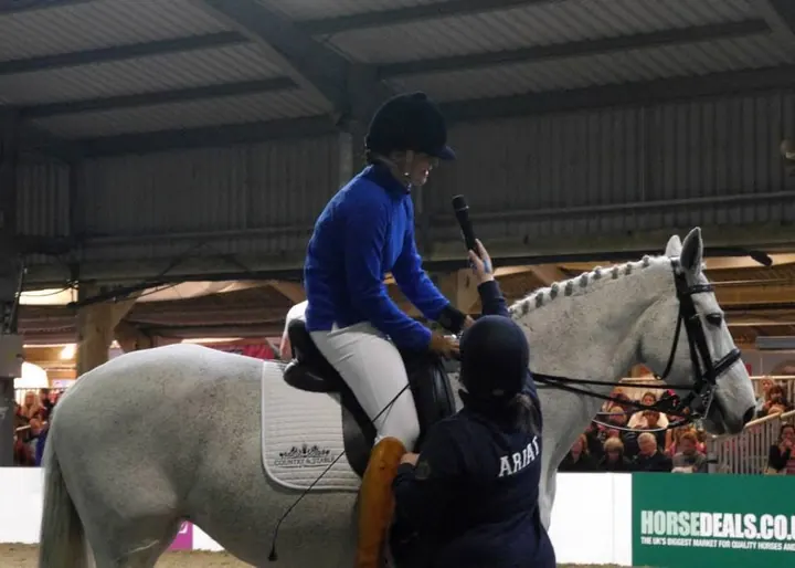 Amanda on a grey horse in an indoor arena, talking to the audience through a microphone