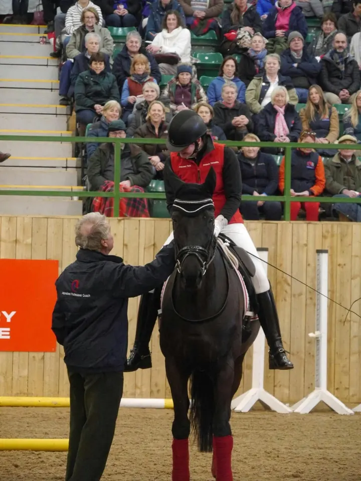 Amanda on a black horse in front of packed stands at a ridden demonstration, with a coach beside her