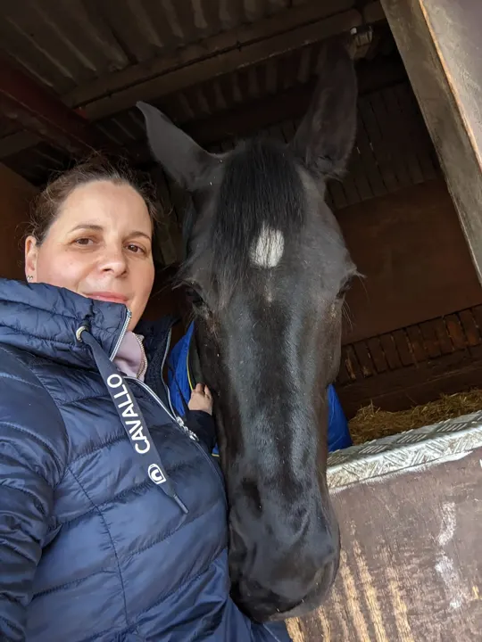 Amanda in a navy jacket with a black horse looking over the stable door